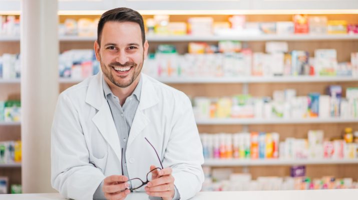 Portrait of a cheerful handsome pharmacist leaning on counter at drugstore.